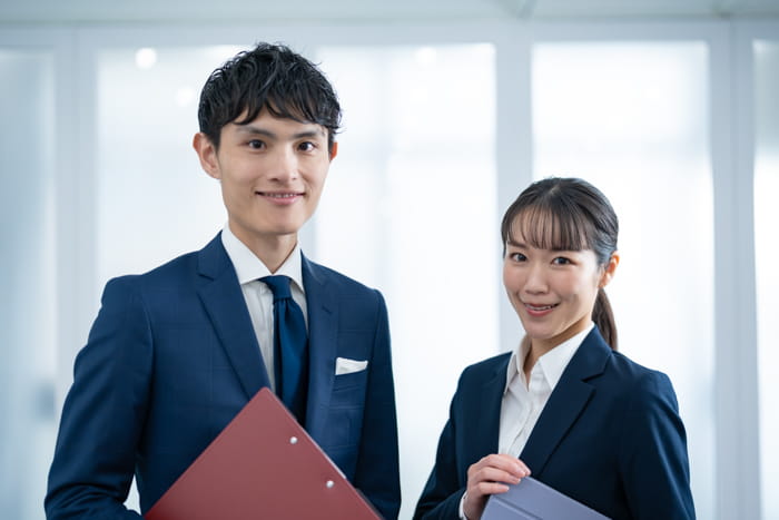 スーツ姿の男性と女性がオフィスで微笑みながら資料を持っている様子。